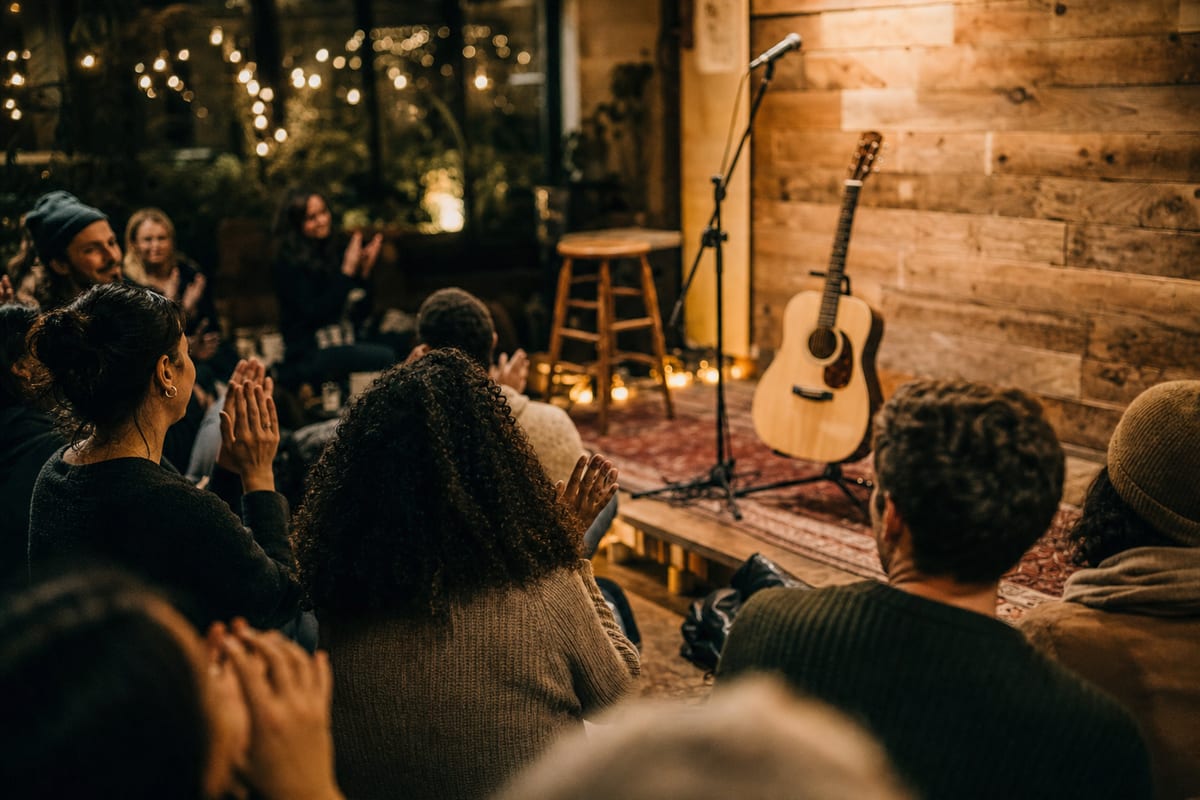 A small engaged crowd claps at an intimate live show while an acoustic guitar rests on a stand on a warmly lit stage.