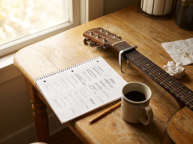 Country songwriting notebook, guitar neck, pencil, and coffee on a kitchen table.