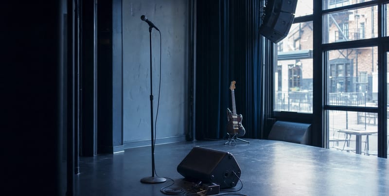 An empty small venue stage with a microphone, wedge monitor, cable, and a guitar resting on a stand in cool blue-grey light, no readable text.