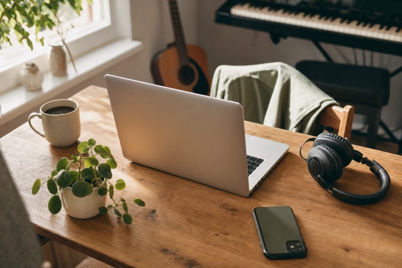 Editorial photo: a musician updating their artist profile on a laptop at a kitchen table, soft morning light, phone and coffee nearby, no readable text
