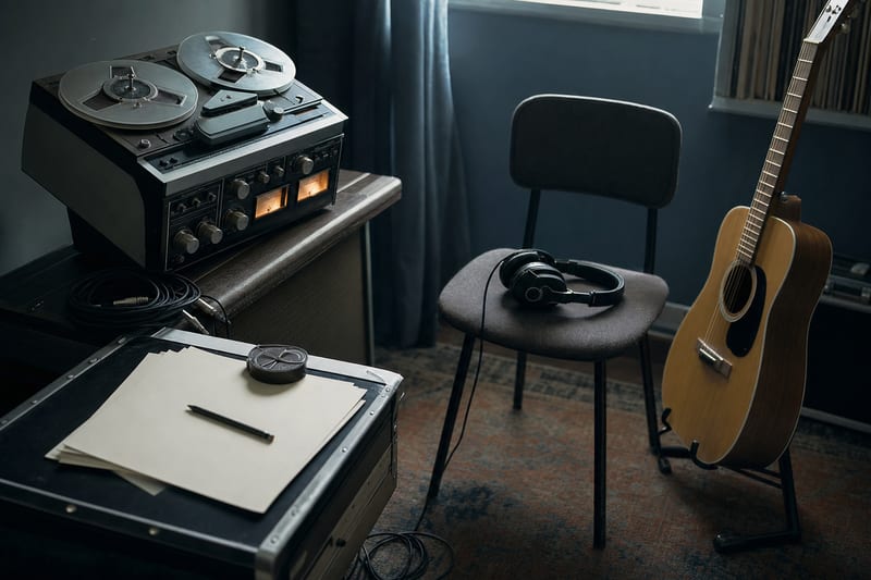 Cool blue studio corner with a reel, blank track notes, headphones, and an acoustic guitar resting safely beside a chair.