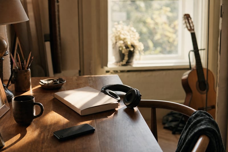 Warm editorial desk with a blank notebook, headphones, a phone turned away, and a guitar resting on a stand near a window.