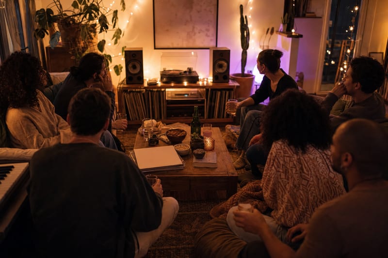 Editorial photo: a small group of friends listening to music together on a phone and speaker in a living room, warm evening light, no readable text