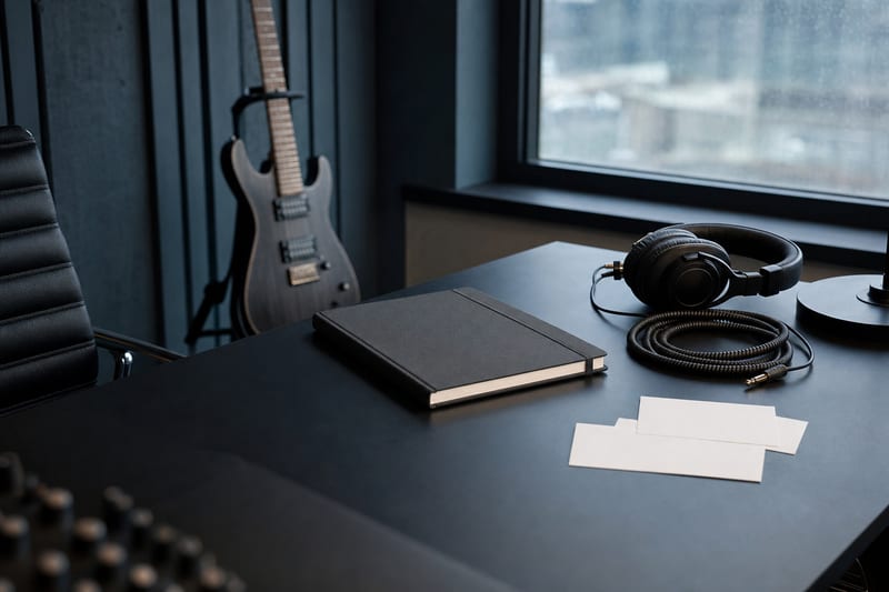 Cool blue-gray studio table with a closed charcoal notebook, blank level cards, headphones, cable, and a small intact guitar resting beside an audio interface.