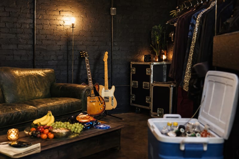 A backstage green room with a worn leather sofa, a snack and fruit tray, a cooler of drinks, and two electric guitars resting on stands against a brick wall.